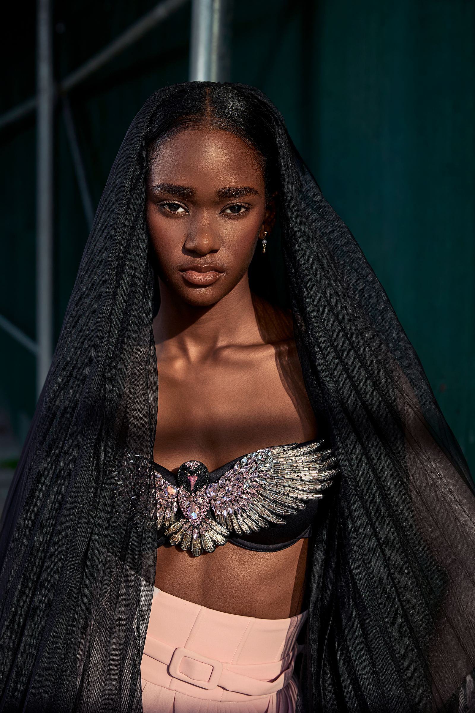 Editorial portrait by RJ Artistry: a model in a sheer black veil and a crystal-winged top looks straight into the camera, a dark teal New York street behind her